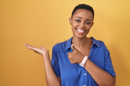 Téléchargez les photos : Femme afro-américaine debout sur fond jaune montrant la main de la paume et faisant un geste correct avec les pouces levés, souriant heureux et joyeux - en image libre de droit