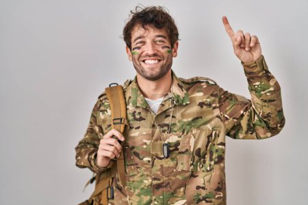 Foto de Joven hispano vistiendo uniforme camuflaje del ejército sonriendo asombrado y sorprendido y señalando con los dedos y los brazos levantados. - Imagen libre de derechos