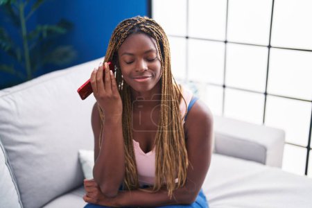 Téléchargez les photos : Femme afro-américaine écoutant un message vocal par smartphone assis sur le canapé à la maison - en image libre de droit
