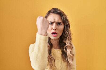 Photo for Young caucasian woman standing over yellow background angry and mad raising fist frustrated and furious while shouting with anger. rage and aggressive concept. - Royalty Free Image
