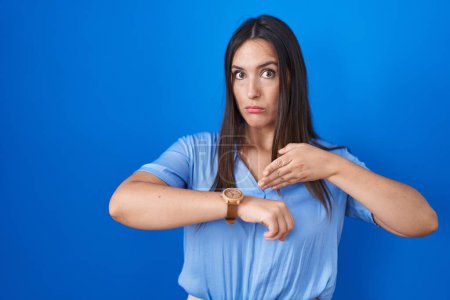 Téléchargez les photos : Jeune femme brune debout sur fond bleu pressé pointant pour regarder le temps, l'impatience, bouleversé et en colère pour retard de délai - en image libre de droit