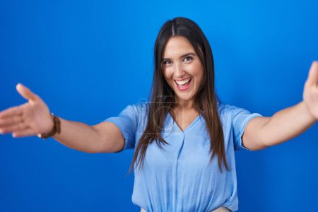 Téléchargez les photos : Jeune femme brune debout sur fond bleu regardant la caméra souriant à bras ouverts pour se faire câliner. expression joyeuse embrassant le bonheur. - en image libre de droit