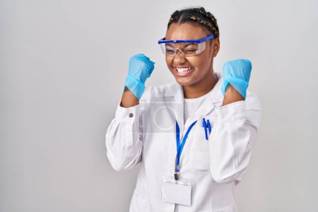 Téléchargez les photos : Femme afro-américaine avec des tresses portant robe de scientifique très heureux et excité de faire geste gagnant avec les bras levés, souriant et criant pour le succès. concept de célébration. - en image libre de droit