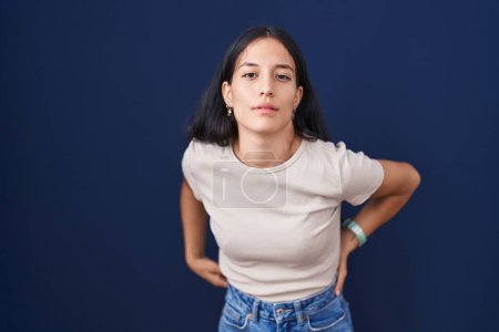 Téléchargez les photos : Jeune femme hispanique debout sur fond bleu souffrant de maux de dos, toucher le dos avec la main, douleur musculaire - en image libre de droit