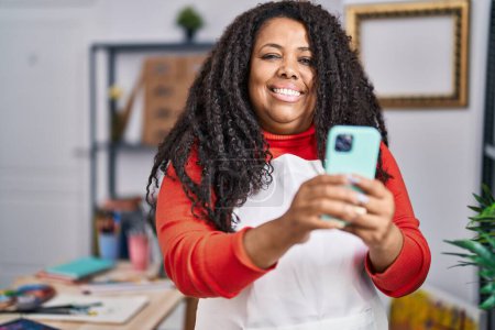 Téléchargez les photos : Femme artiste afro-américaine souriant confiant en utilisant smartphone au studio d'art - en image libre de droit