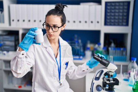 Téléchargez les photos : Jeune scientifique caucasienne utilisant un microscope tenant une bouteille de pilules au laboratoire - en image libre de droit