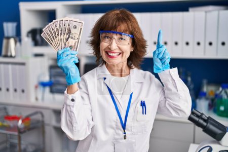 Photo for Middle age woman working at scientist laboratory holding money surprised with an idea or question pointing finger with happy face, number one - Royalty Free Image