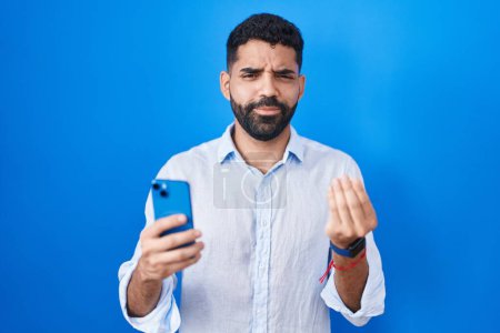 Téléchargez les photos : Homme hispanique avec barbe en utilisant le message de dactylographie smartphone faire geste d'argent avec les mains, demandant le paiement du salaire, entreprise millionnaire - en image libre de droit