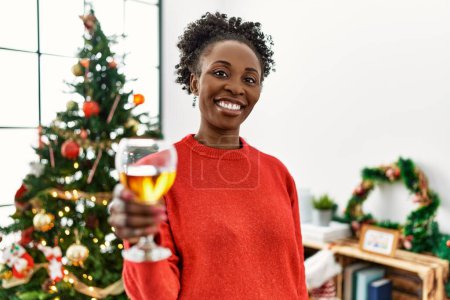 Téléchargez les photos : Femme afro-américaine buvant du champagne debout près de l'arbre de Noël à la maison - en image libre de droit