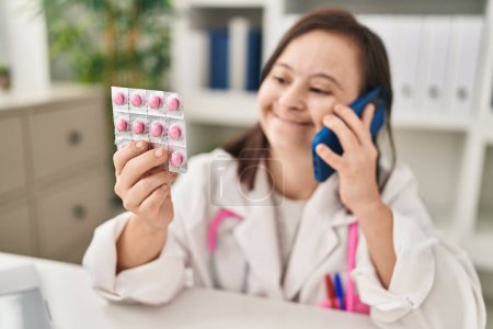 Foto de Síndrome de Down mujer que usa uniforme médico hablando en el teléfono inteligente que contiene píldoras anticonceptivas en la clínica - Imagen libre de derechos