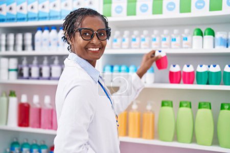 Foto de Farmacéutica afroamericana de mediana edad sonriendo confiada sosteniendo pasta de dientes en estanterías en la farmacia - Imagen libre de derechos