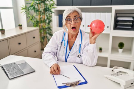 Photo for Middle age woman with grey hair wearing doctor uniform holding balloon scared and amazed with open mouth for surprise, disbelief face - Royalty Free Image