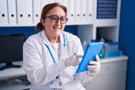 Téléchargez les photos : Femme scientifique senior souriant confiant en utilisant le pavé tactile au laboratoire - en image libre de droit