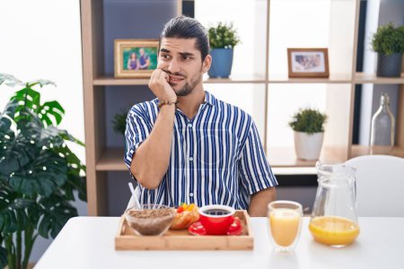 Photo for Hispanic man with long hair sitting on the table having breakfast looking stressed and nervous with hands on mouth biting nails. anxiety problem. - Royalty Free Image