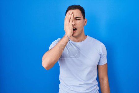 Photo for Young hispanic man standing over blue background yawning tired covering half face, eye and mouth with hand. face hurts in pain. - Royalty Free Image