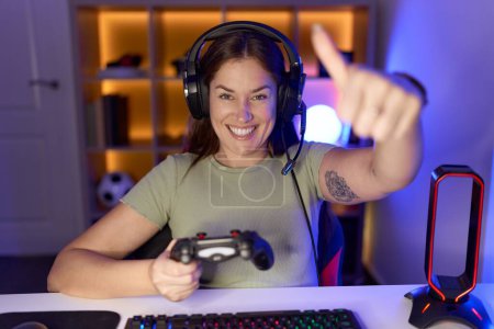 Photo for Beautiful brunette woman playing video games wearing headphones approving doing positive gesture with hand, thumbs up smiling and happy for success. winner gesture. - Royalty Free Image