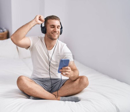 Téléchargez les photos : Young caucasian man dancing and listening to music sititng on bed at bedroom - en image libre de droit