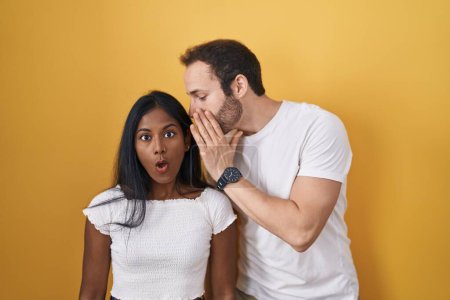 Téléchargez les photos : Couple Interracial debout sur fond jaune main sur bouche racontant rumeur secrète, chuchotant conversation malicieuse - en image libre de droit