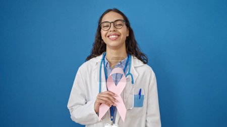 Foto de Joven hermosa mujer hispana médico sosteniendo la conciencia de cáncer de mama cinta rosa sobre fondo azul aislado - Imagen libre de derechos