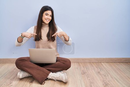 Foto de Mujer morena joven que trabaja usando ordenador portátil sentado en el suelo mirando confiado con sonrisa en la cara, señalándose con los dedos orgullosos y felices. - Imagen libre de derechos
