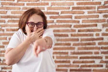Photo for Senior woman with glasses standing over bricks wall laughing at you, pointing finger to the camera with hand over mouth, shame expression - Royalty Free Image