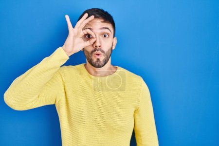 Photo for Hispanic man standing over blue background doing ok gesture shocked with surprised face, eye looking through fingers. unbelieving expression. - Royalty Free Image