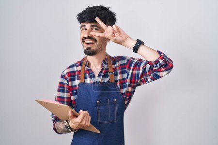 Foto de Joven hombre hispano con barba llevando delantal de camarero sosteniendo portapapeles haciendo símbolo de paz con los dedos sobre la cara, sonriendo alegre mostrando la victoria - Imagen libre de derechos