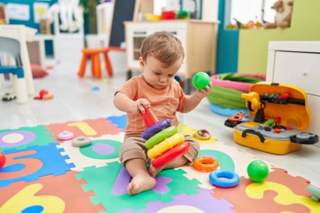 Foto de Adorable niño rubio jugando con aros de juguete y pelota sentado en el suelo en el jardín de infantes - Imagen libre de derechos