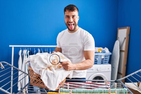 Foto de Hombre hispano guapo sosteniendo lupa buscando mancha en la ropa sacando la lengua feliz con expresión divertida. - Imagen libre de derechos