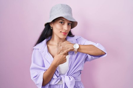 Téléchargez les photos : Jeune femme hispanique debout sur fond rose portant chapeau à la hâte pointant vers regarder le temps, l'impatience, regardant la caméra avec une expression détendue - en image libre de droit