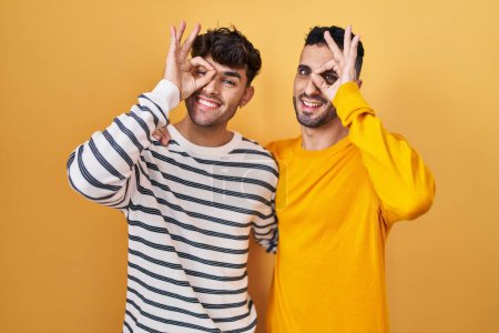 Téléchargez les photos : Jeune couple gay hispanique debout sur fond jaune faisant un geste correct avec la main souriante, les yeux regardant à travers les doigts avec un visage heureux. - en image libre de droit