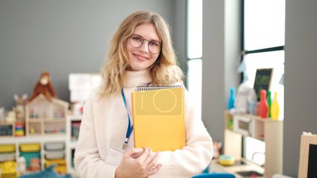 Photo for Young blonde woman preschool teacher smiling confident holding books at kindergarten - Royalty Free Image