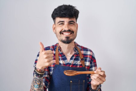 Téléchargez les photos : Jeune homme hispanique avec barbe portant tablier dégustation de nourriture tenant cuillère en bois souriant heureux et positif, pouce levé faire excellent et signe d'approbation - en image libre de droit