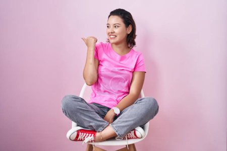 Téléchargez les photos : Jeune femme hispanique assise sur une chaise sur fond rose souriant avec le visage heureux regardant et pointant vers le côté avec le pouce vers le haut. - en image libre de droit
