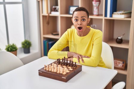 Foto de Mujer afroamericana jugando al ajedrez sentada en la mesa asustada y sorprendida con la boca abierta por sorpresa, cara de incredulidad - Imagen libre de derechos