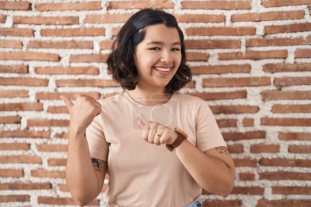 Foto de Mujer hispana joven de pie sobre la pared de ladrillos apuntando hacia atrás con la mano y los pulgares hacia arriba, sonriendo confiada - Imagen libre de derechos