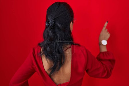 Téléchargez les photos : Asiatique jeune femme debout sur fond rouge posant vers l'arrière pointant vers l'avant avec la main du doigt - en image libre de droit