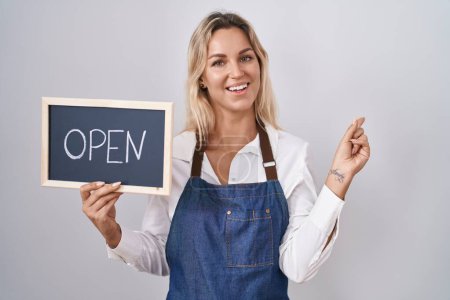 Photo for Young blonde woman wearing apron holding blackboard with open word smiling happy pointing with hand and finger to the side - Royalty Free Image