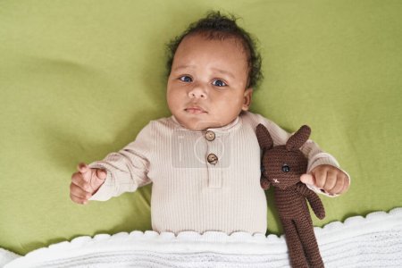 Téléchargez les photos : Bébé afro-américain se relaxant sur le lit avec poupée dans la chambre - en image libre de droit
