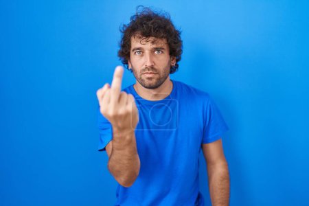 Foto de Hispanic young man standing over blue background showing middle finger, impolite and rude fuck off expression - Imagen libre de derechos