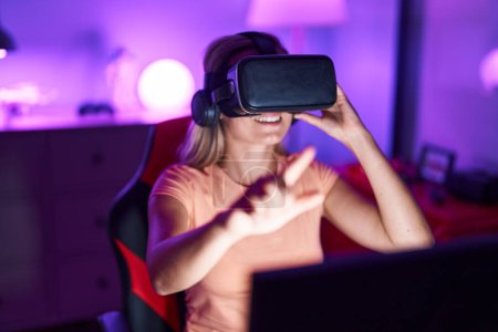 Téléchargez les photos : Jeune femme blonde streamer jouer à un jeu vidéo en utilisant des lunettes de réalité virtuelle à la salle de jeu - en image libre de droit
