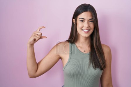 Foto de Hispanic woman standing over pink background smiling and confident gesturing with hand doing small size sign with fingers looking and the camera. measure concept. - Imagen libre de derechos