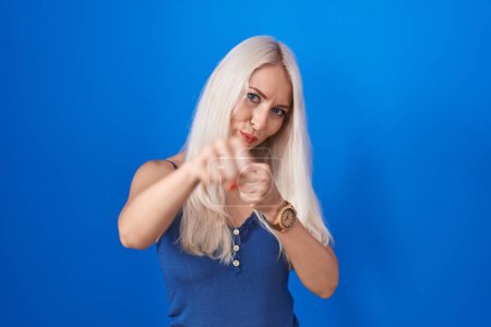 Téléchargez les photos : Femme caucasienne debout sur fond bleu coup de poing poing pour se battre, attaque agressive et en colère, menace et violence - en image libre de droit
