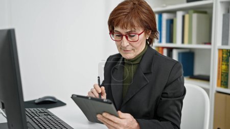 Téléchargez les photos : Mature hispanique femme professeur à l'aide de l'ordinateur prenant des notes sur le pavé tactile à la bibliothèque de l'université - en image libre de droit