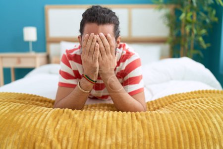 Téléchargez les photos : Jeune homme hispanique couché sur le lit couvrant les yeux avec les mains à la chambre - en image libre de droit