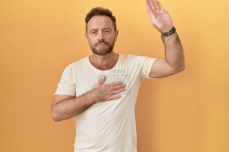 Photo for Middle age man with beard standing over yellow background swearing with hand on chest and open palm, making a loyalty promise oath - Royalty Free Image