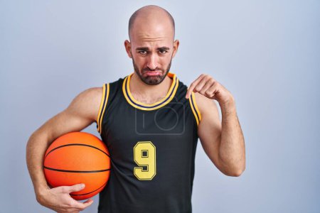 Téléchargez les photos : Jeune homme chauve avec barbe portant l'uniforme de basket tenant la balle pointant vers le bas regardant triste et contrarié, indiquant la direction avec les doigts, malheureux et déprimé. - en image libre de droit