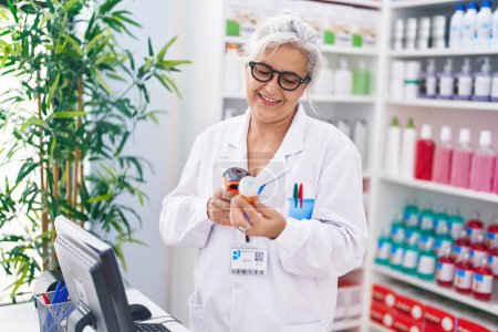 Téléchargez les photos : Moyen âge femme aux cheveux gris pharmacien numérisation pilules bouteille à la pharmacie - en image libre de droit