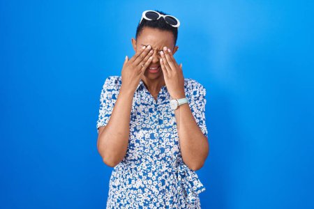 Foto de Mujer afroamericana de pie sobre fondo azul frotando los ojos para la fatiga y el dolor de cabeza, la expresión somnolienta y cansada. problema de visión - Imagen libre de derechos