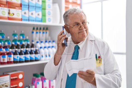 Téléchargez les photos : Middle age grey-haired man pharmacist talking on smartphone reading prescription at pharmacy - en image libre de droit